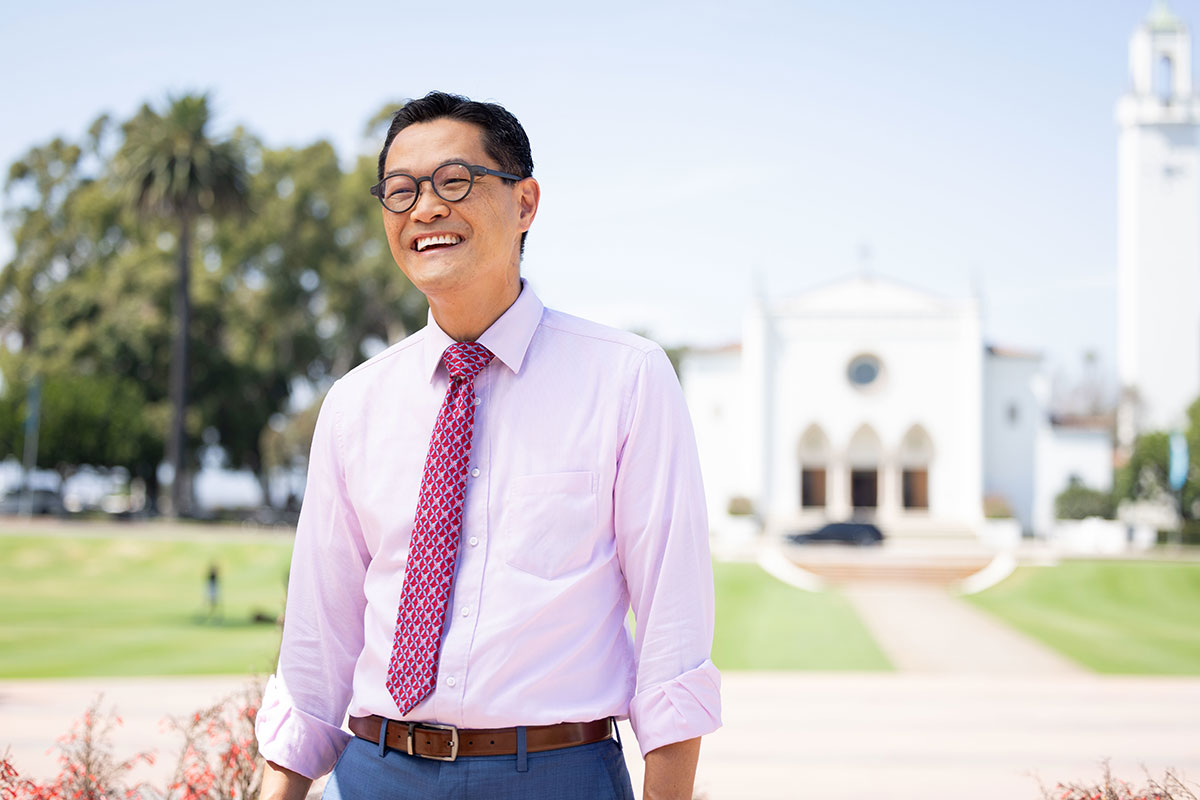President Thomas Poon smiling while standing in front of Sacred Heart Chapel and Sunken Garden
